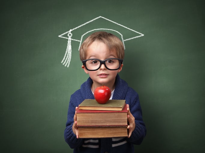 Young boy wearing glasses holding books with an apple on top and a chalk drawn graduation cap